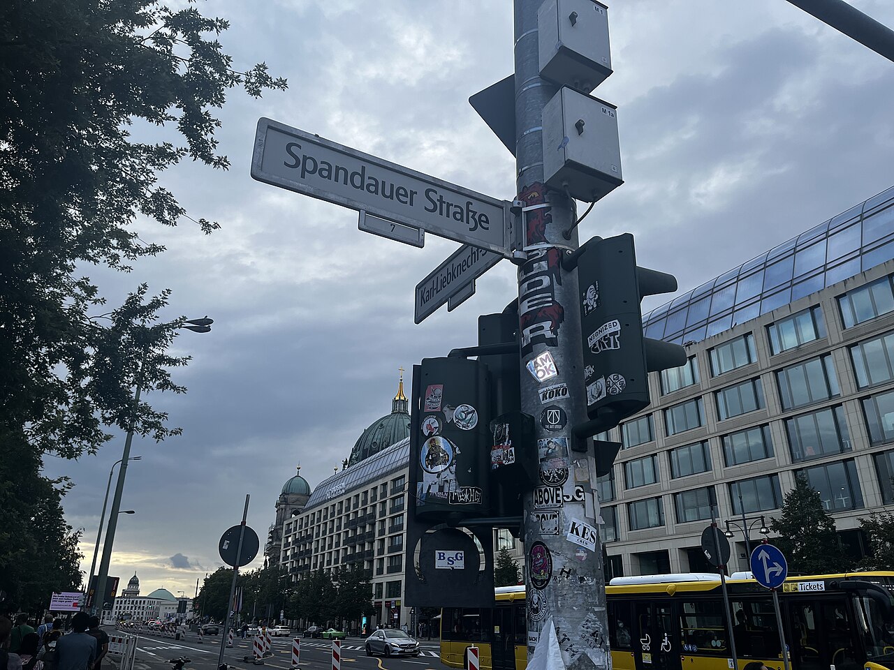 Street sign reading Spandauer Straße on a pole covered in stickers, with a yellow bus and large buildings in Berlin behind it photo_0